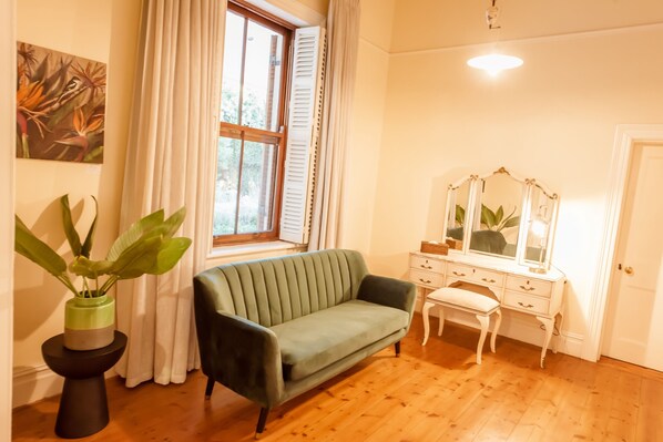 Una habitación con sofá verde, cómoda blanca con espejo, planta en maceta y ventana con cortinas blancas.