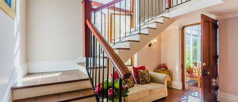 A staircase with wooden railings leading to an upper floor, a beige sofa with colorful cushions, and a patterned rug on the floor.