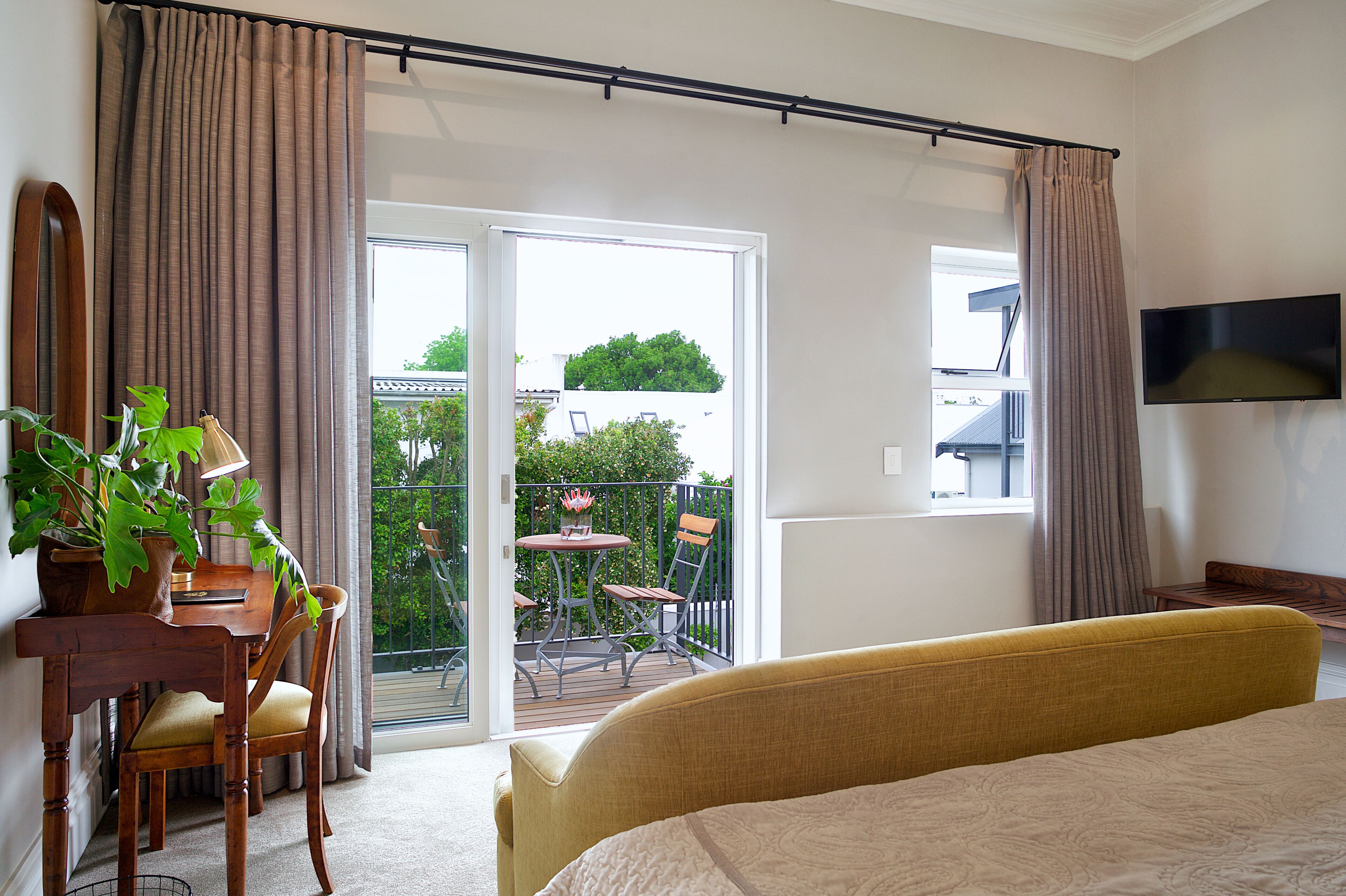 Une chambre d’hôtel comprenant un lit, un bureau avec une chaise, une plante en pot, un balcon avec une table et des chaises, et un téléviseur fixé au mur.
