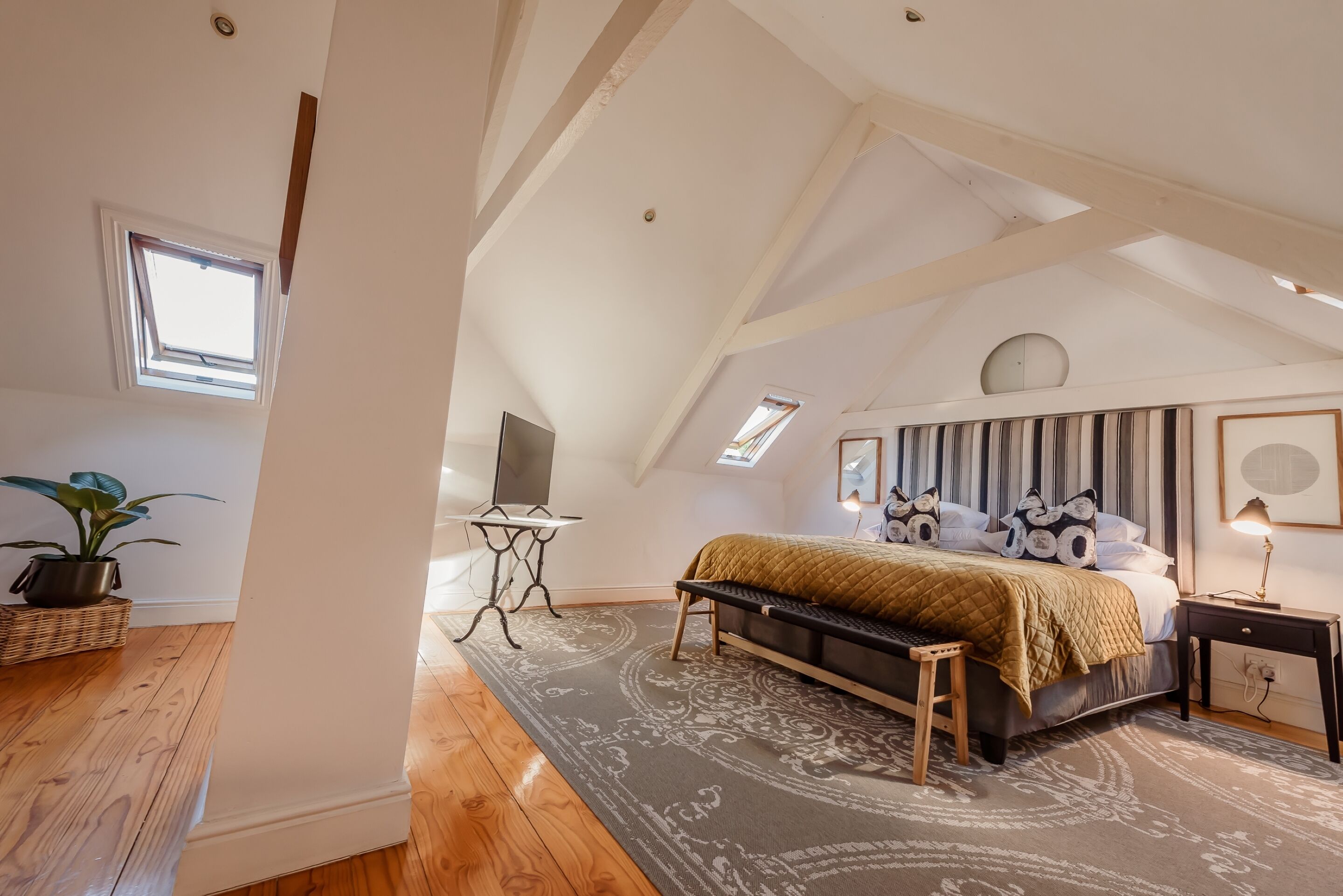 A bedroom with a sloped ceiling, a bed with a patterned bedspread, a bench, a small table, and a potted plant.