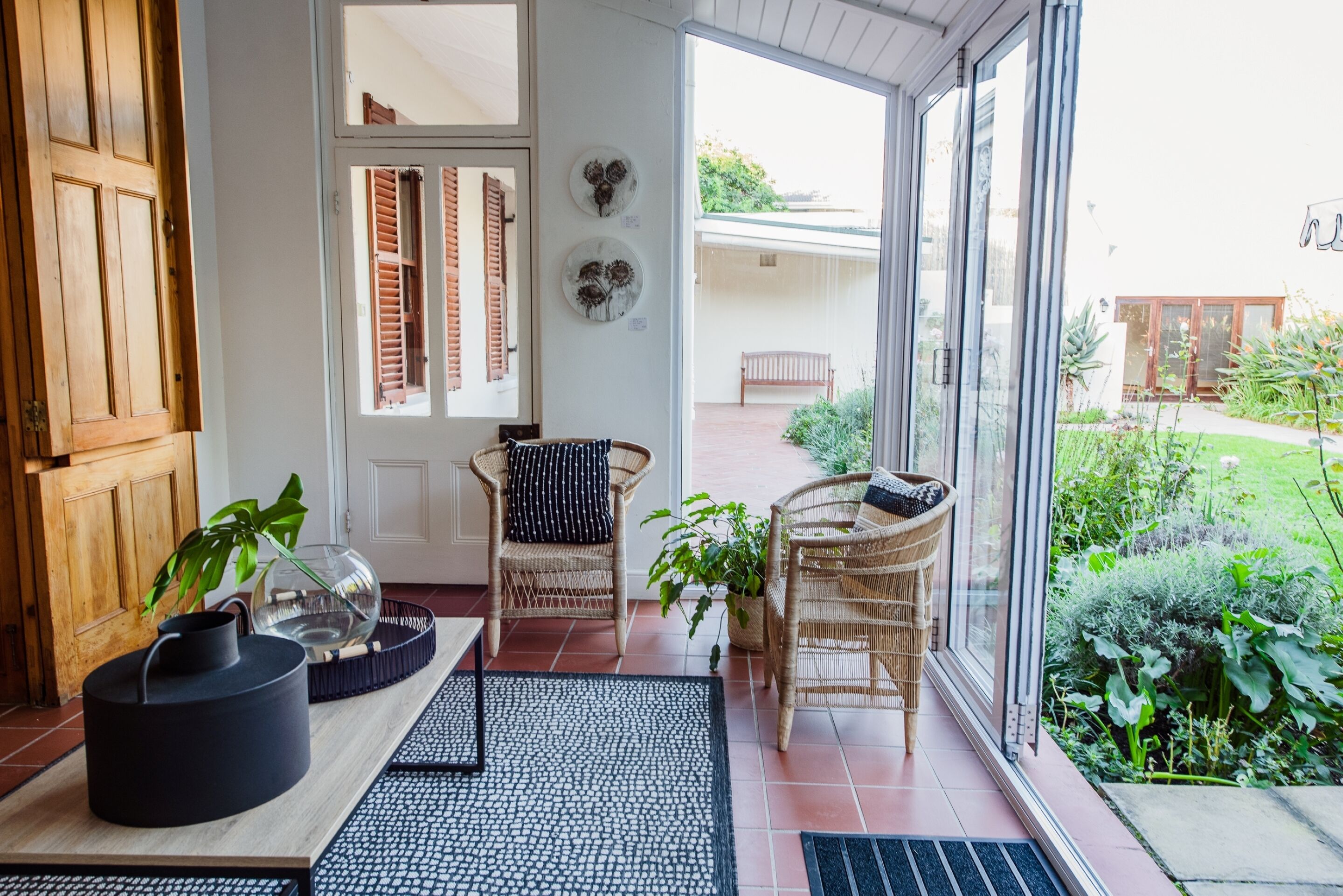 Una habitación iluminada por el sol con puerta de madera, sillas de mimbre, puerta de cristal que da al exterior y una planta en maceta.