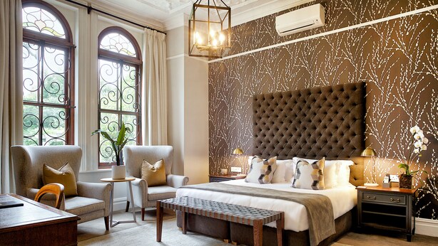 Een slaapkamer met een groot bed, twee fauteuils, een houten bankje, een bureau en een nachtkastje met een lamp.