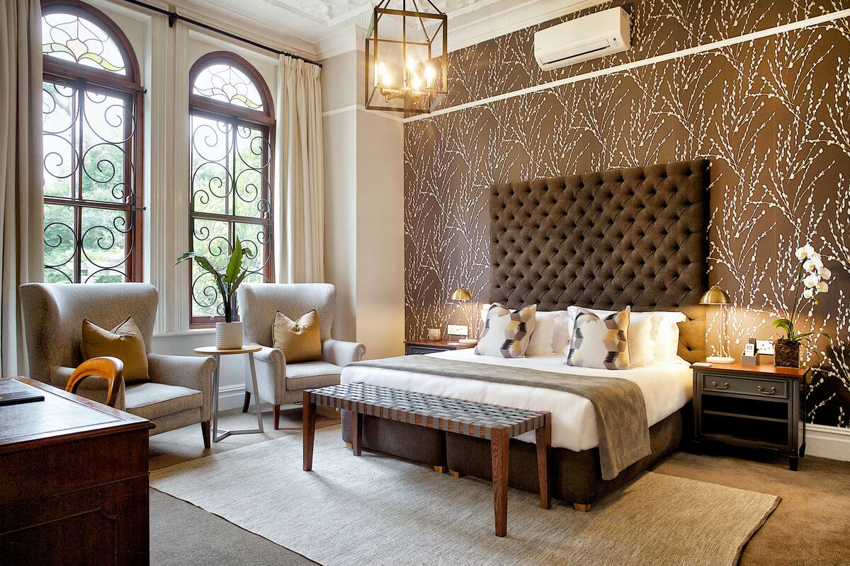 Een slaapkamer met een groot bed, twee fauteuils, een houten bankje, een bureau en een nachtkastje met een lamp.