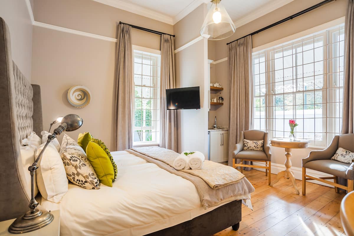 Een goed verlichte slaapkamer met een groot bed, een nachtkastje met een lamp, twee stoelen, een kleine tafel met een vaas, en een televisie aan de muur.