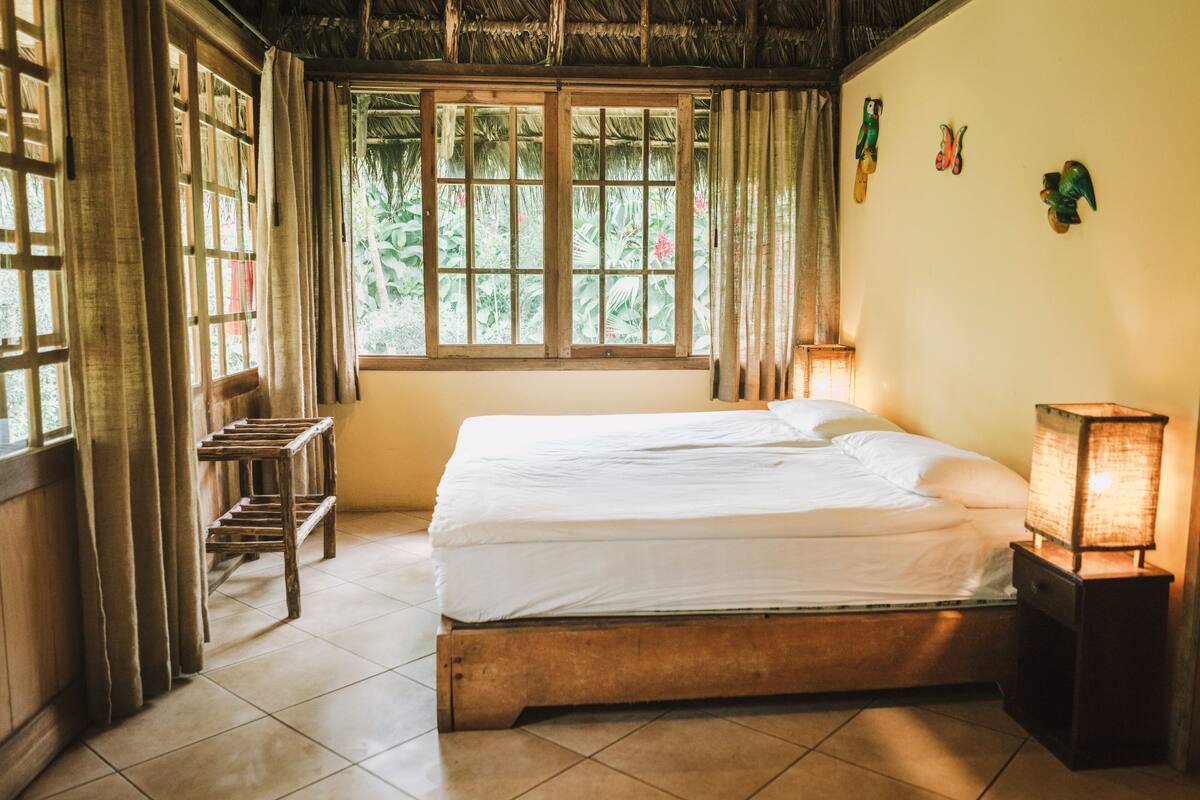 A room with a bed, a nightstand with a lamp, a window with curtains, and a wall with a bird sculpture.