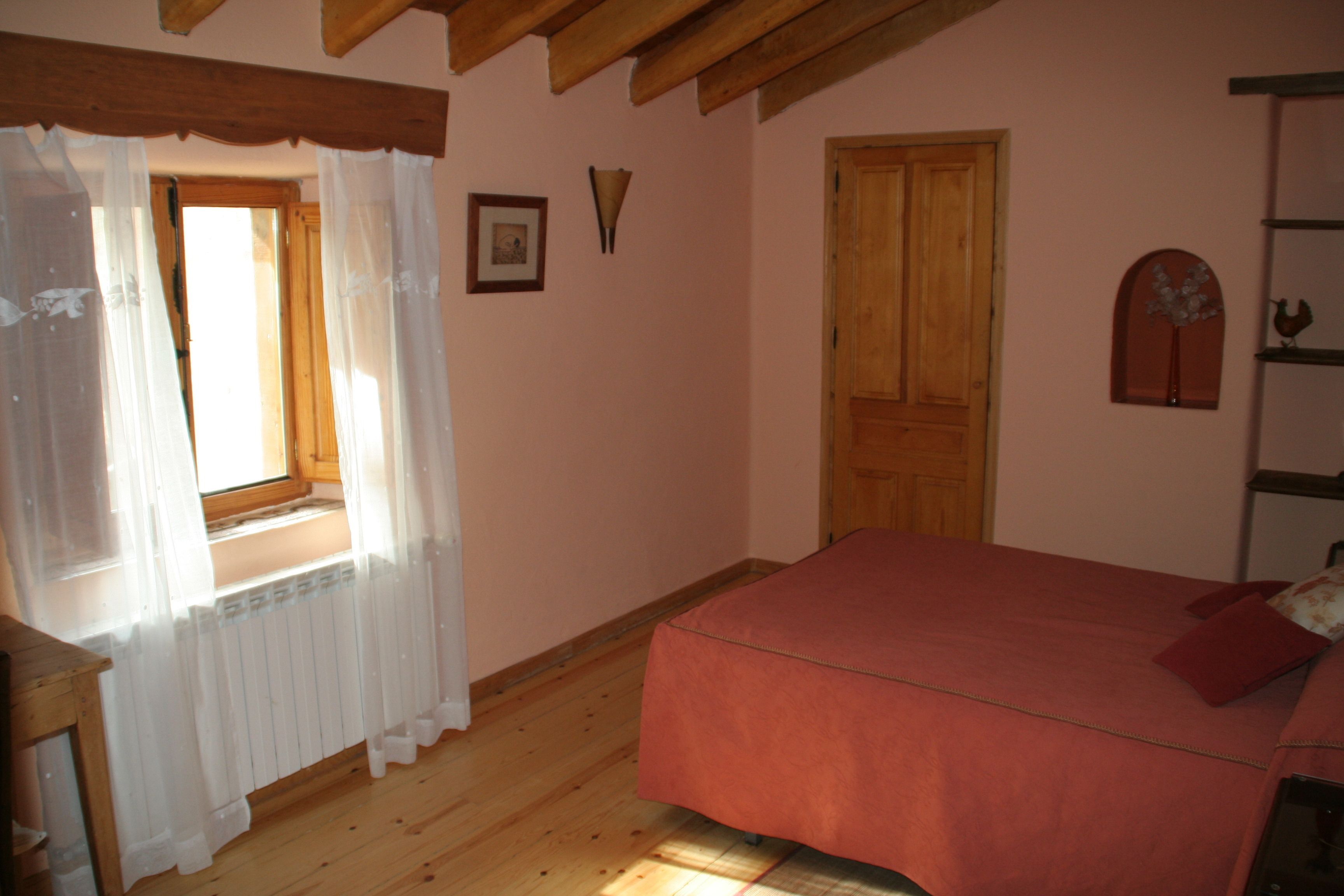 Un dormitorio con suelo de madera, una cama roja, una ventana con cortinas traslúcidas y una puerta de madera.