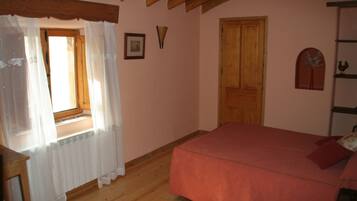 Un dormitorio con suelo de madera, una cama roja, una ventana con cortinas traslúcidas y una puerta de madera.