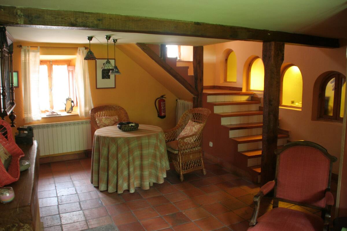 Una habitación acogedora con una mesa redonda, sillas de mimbre, una chimenea y una escalera con ventanales arqueados.