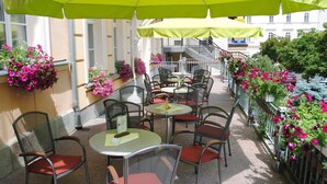 An outdoor café with round tables and chairs, covered by green umbrellas, and surrounded by flower boxes with pink flowers. - Venus (Karlovy Vary)