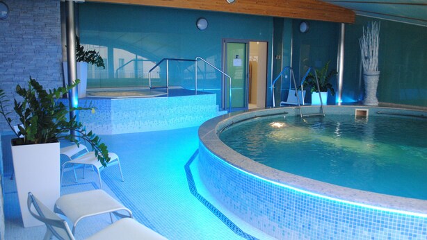 An indoor swimming pool with clear blue water, tiled edges, and a wooden ceiling. There are lounge chairs and potted plants around the pool area. - Venus (Karlovy Vary)