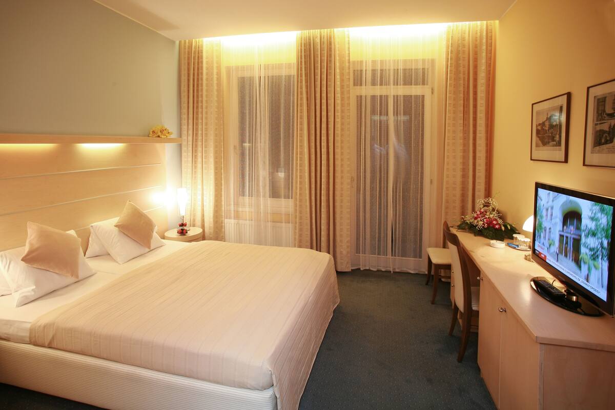Ein Hotelzimmer mit einem großen Bett, Nachttischen, einem Schreibtisch mit Stuhl, einem Fernseher und Vorhängen.