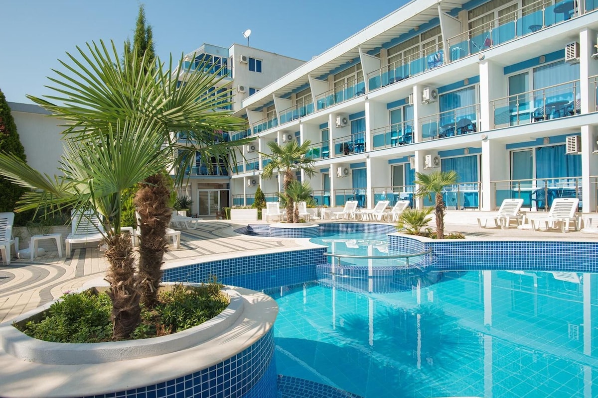 A hotel pool area with multiple levels of balconies, lounge chairs, and palm trees.