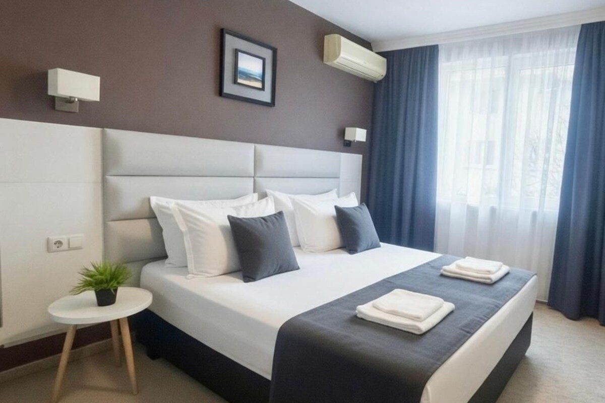 A neatly made bed with a headboard, a small round table with a plant, and a window with curtains.