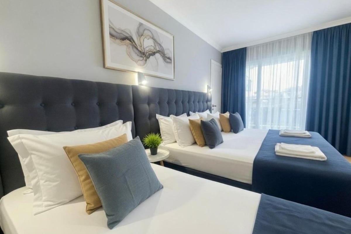 A hotel room with two beds, a large headboard, a wall-mounted artwork, and a window with curtains.