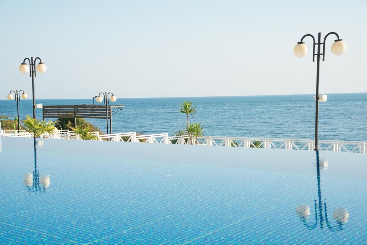 An infinity pool with a clear view of the ocean, a wooden deck, and two lamp posts.