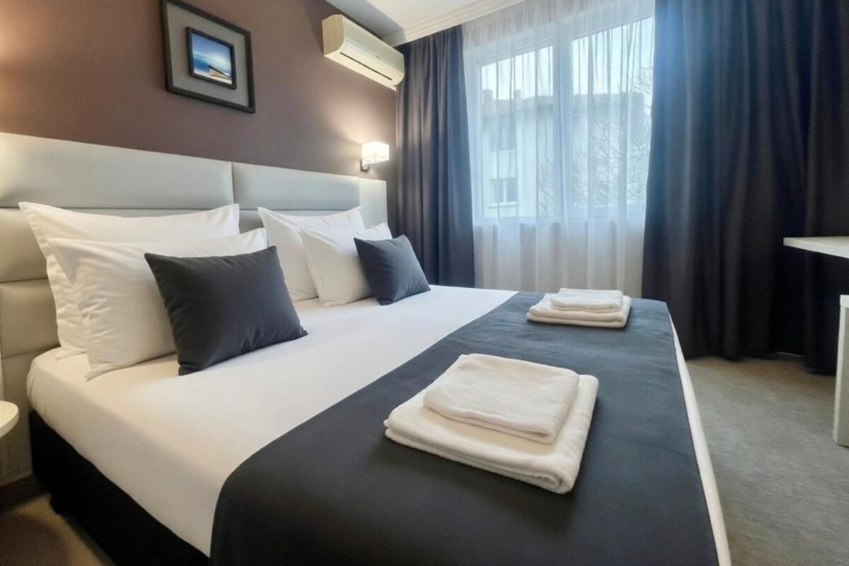 A hotel room with a large bed, two bedside tables, a wall-mounted air conditioner, and a window with curtains.