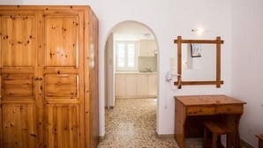 Una habitación con un armario de madera, un escritorio con cajones, un espejo y un arco que da a una cocina.