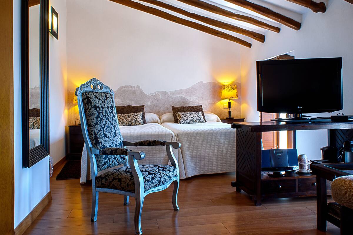 A bedroom with a bed, two bedside tables, a chair, a desk with a TV, and a lamp.