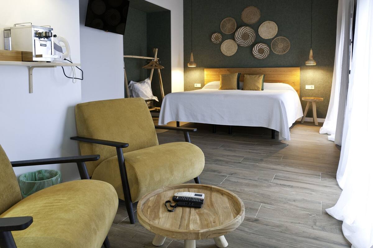 A hotel room with a bed, two chairs, a coffee machine, and a small table with a remote control.