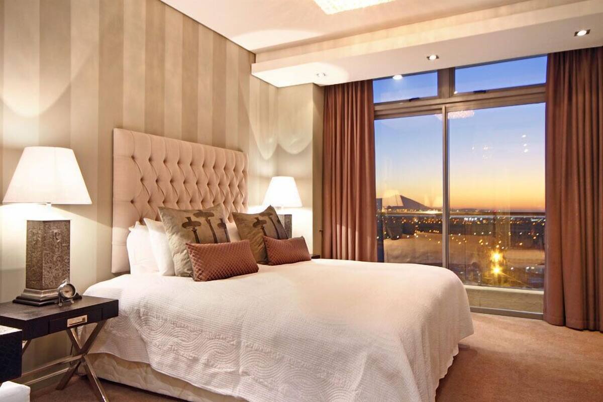 Ein geräumiges Schlafzimmer mit einem großen Bett, zwei Nachttischen und Blick auf eine Stadtlandschaft bei Sonnenuntergang.