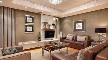 A modern living room with a glass coffee table, leather sofas, a flat-screen TV, framed artwork, and a chandelier.