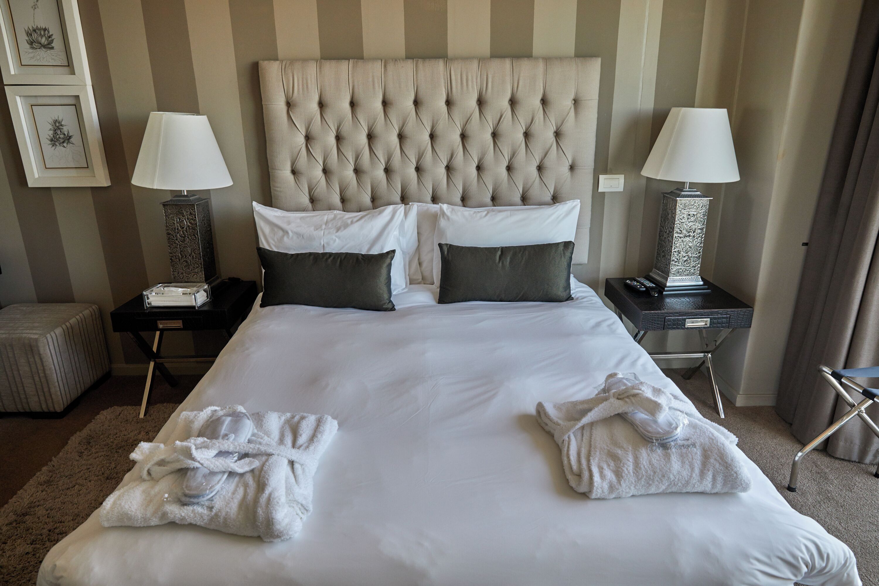A hotel room with a large bed, two bedside tables with lamps, a headboard, and a small ottoman.