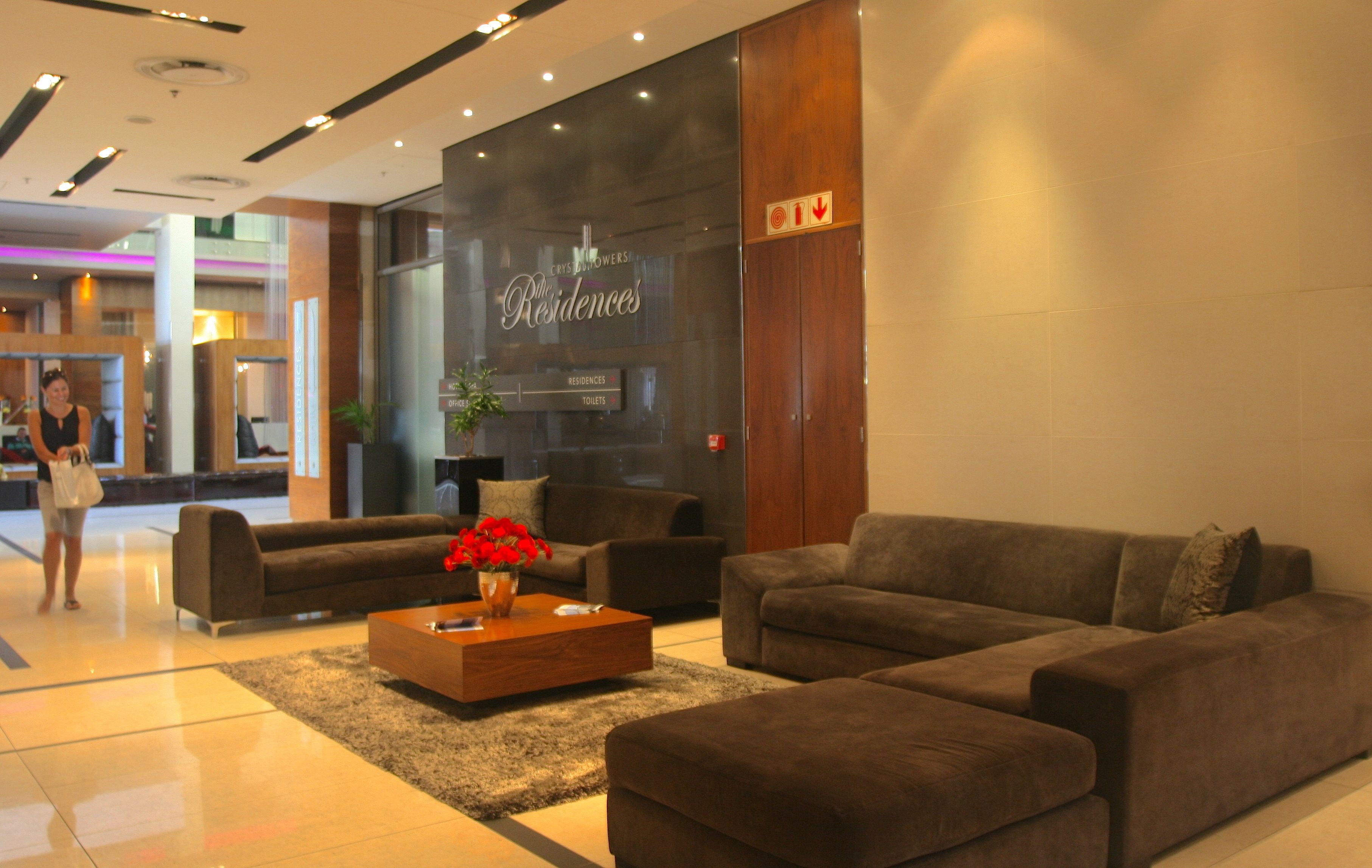 A hotel lobby with a reception desk, a seating area with sofas, a coffee table with flowers, and a sign indicating the location of the bathrooms.