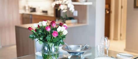 Una mesa de comedor con un florero de vidrio transparente con flores, una bandeja de metal y un juego de cubiertos.