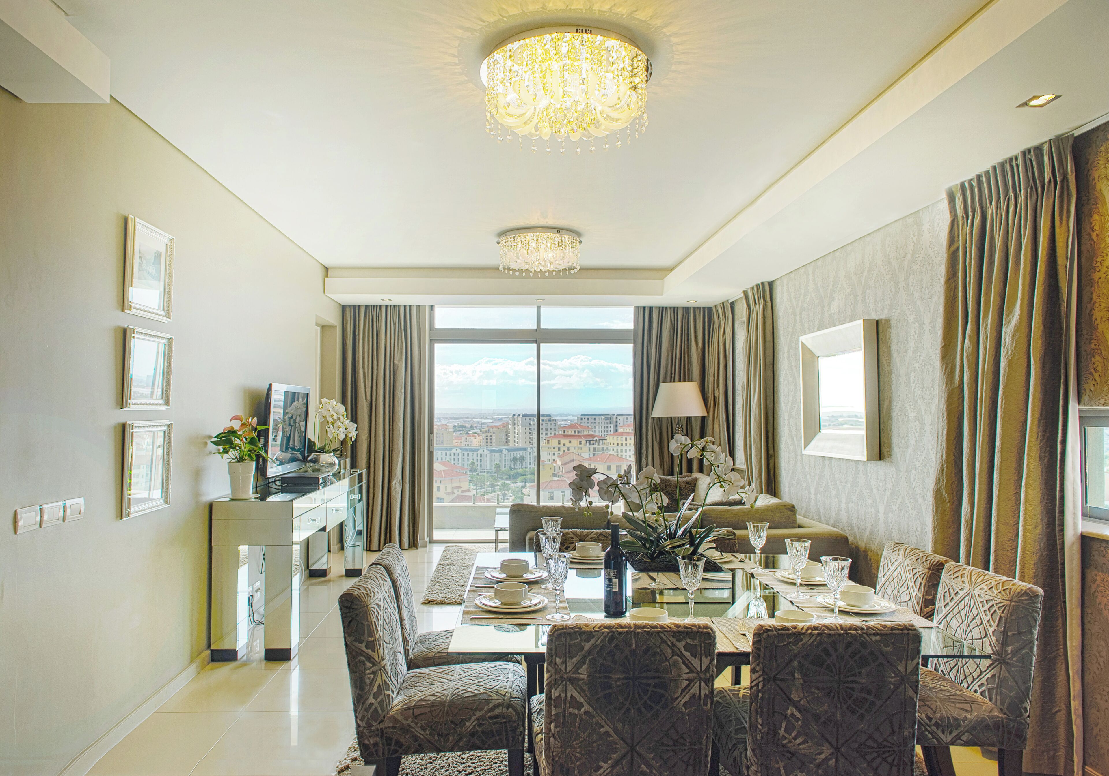 A dining area with a table set for four, a chandelier, and a city view through large windows.