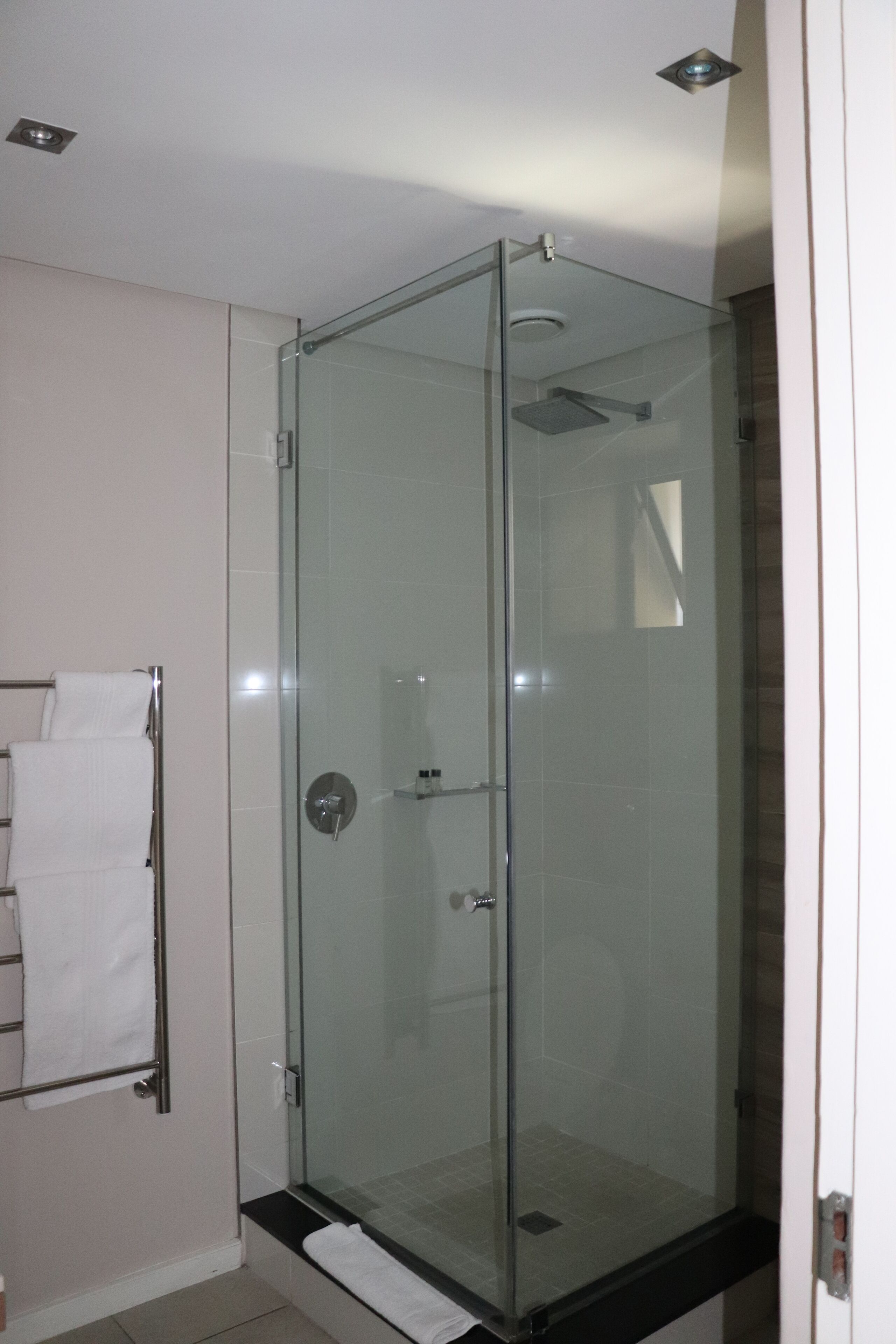 A glass-enclosed shower stall with a white tiled floor, a towel rack, and a small shelf with toiletries.