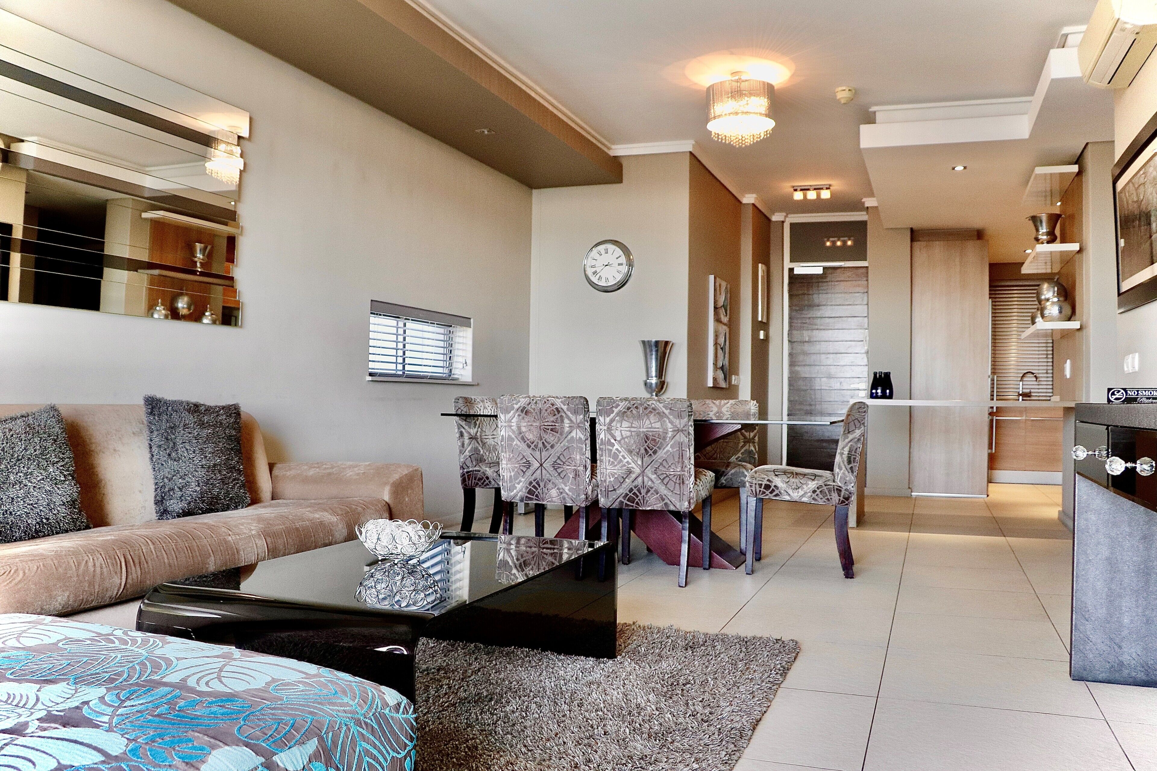 A modern living room with a sofa, coffee table, patterned rug, dining area with chairs, and a kitchen area with a countertop and cabinets.