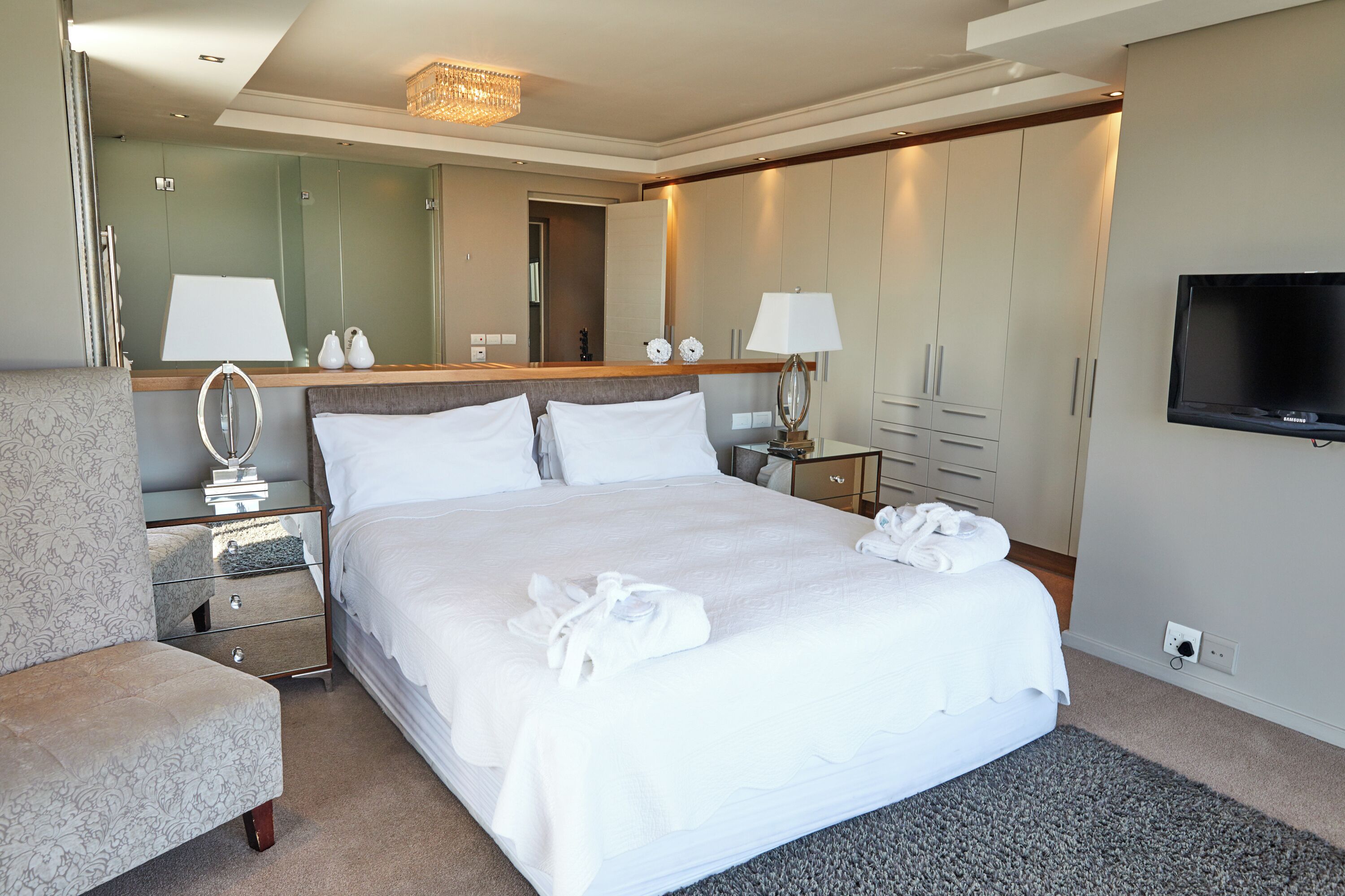 A hotel room with a large bed, a bedside table with lamps, a mirror, a wardrobe, and a TV.