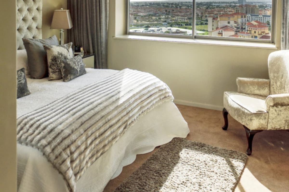 A bedroom with a large bed, a chair, a chandelier, and a view of a cityscape through a large window.