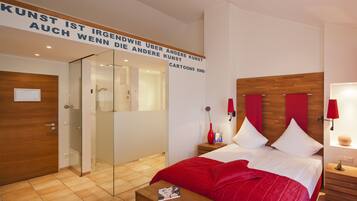 A hotel room with a bed, a nightstand, a bedside table, a mirror, a red wall art, and a shower area.