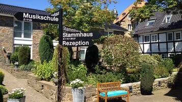 Un patio con un banco de madera, paredes de piedra y plantas en macetas. Se observan letreros que indican 'Musikschule' y 'Stammhaus' con recepción.