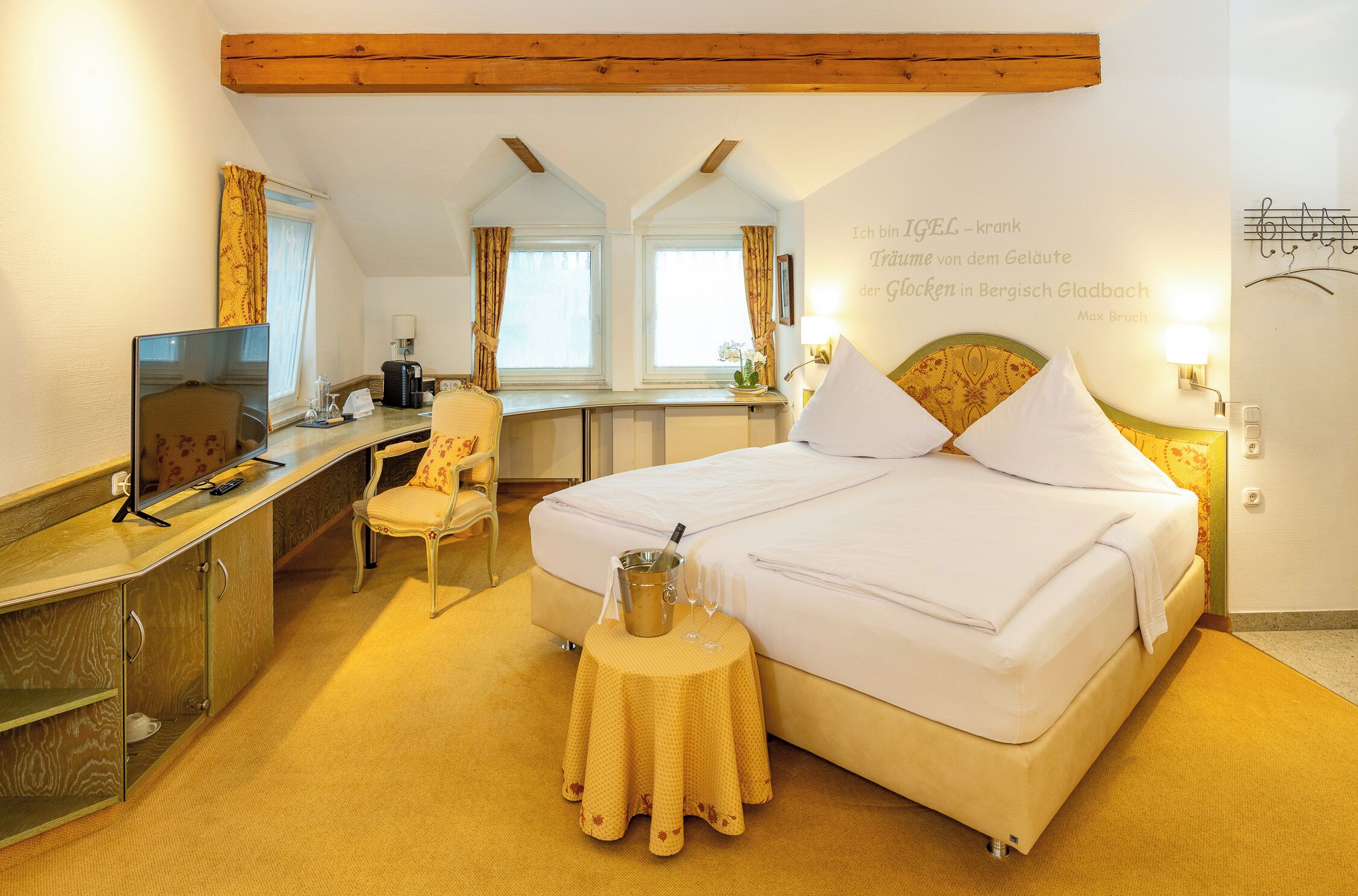 A hotel room with a bed, a desk, a chair, a television, and a small table with a bottle and glasses.