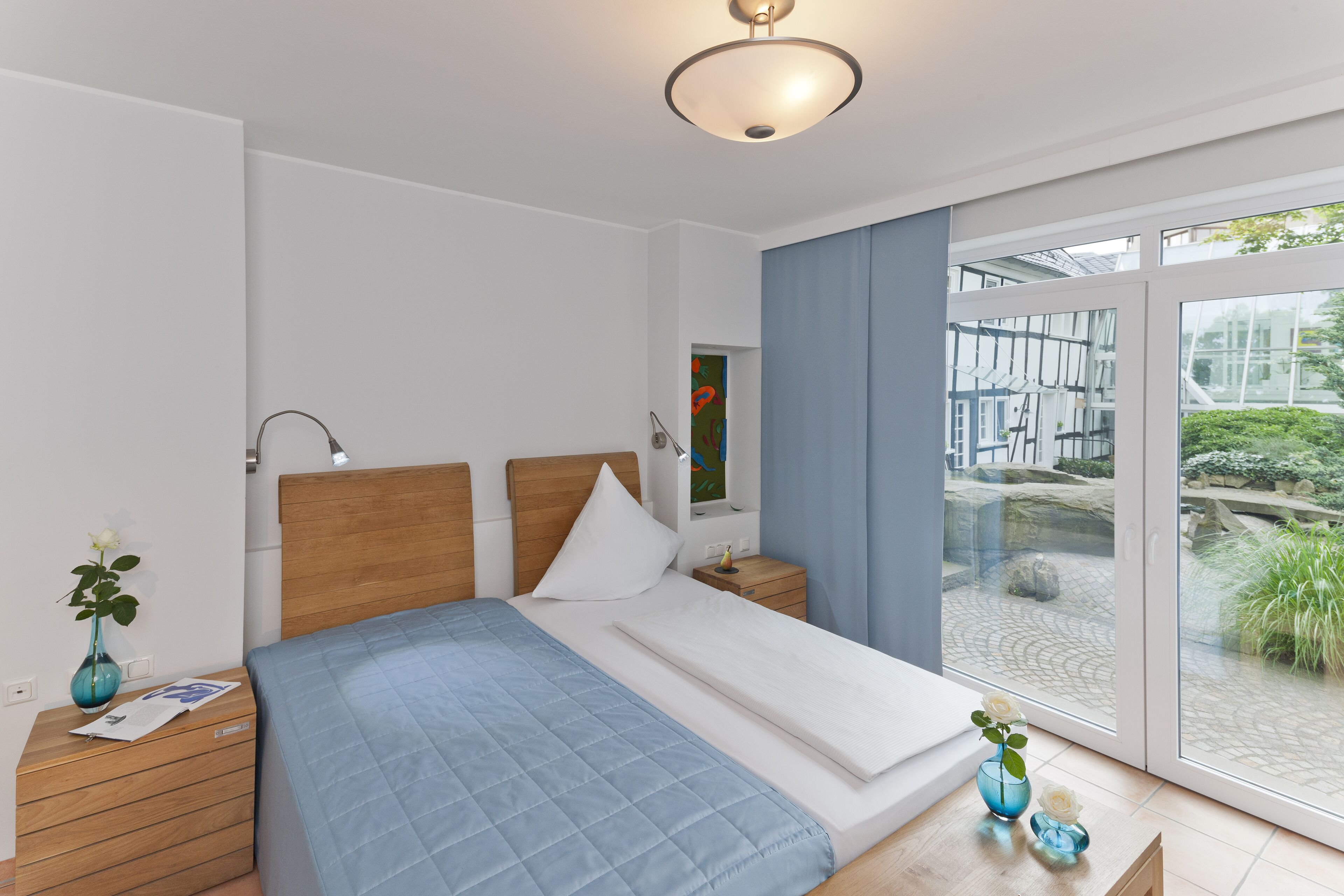 Ein modernes Schlafzimmer mit einem großen Bett, einem Holz-Kopfteil, Nachttischen, einem Beistelltisch, einem Fenster mit Blick auf Grünflächen und einer Deckenleuchte.