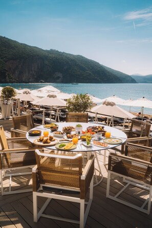 An outdoor dining area with wicker chairs and tables, overlooking a body of water and a mountainous landscape. - Lichnos Beach Hotel & Suites (Parga)