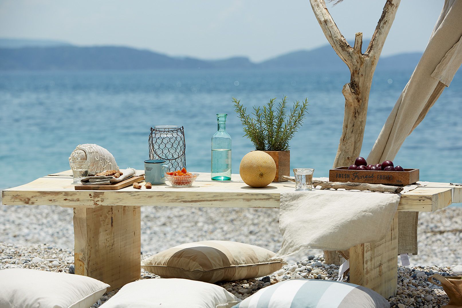 A wooden table set on a pebble beach with a view of the sea and mountains.