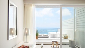 A modern hotel room with a large window offering a view of the sea. There is a dining area with a table, a bowl of fruit, and a lamp. - Lichnos Beach Hotel & Suites (Parga)