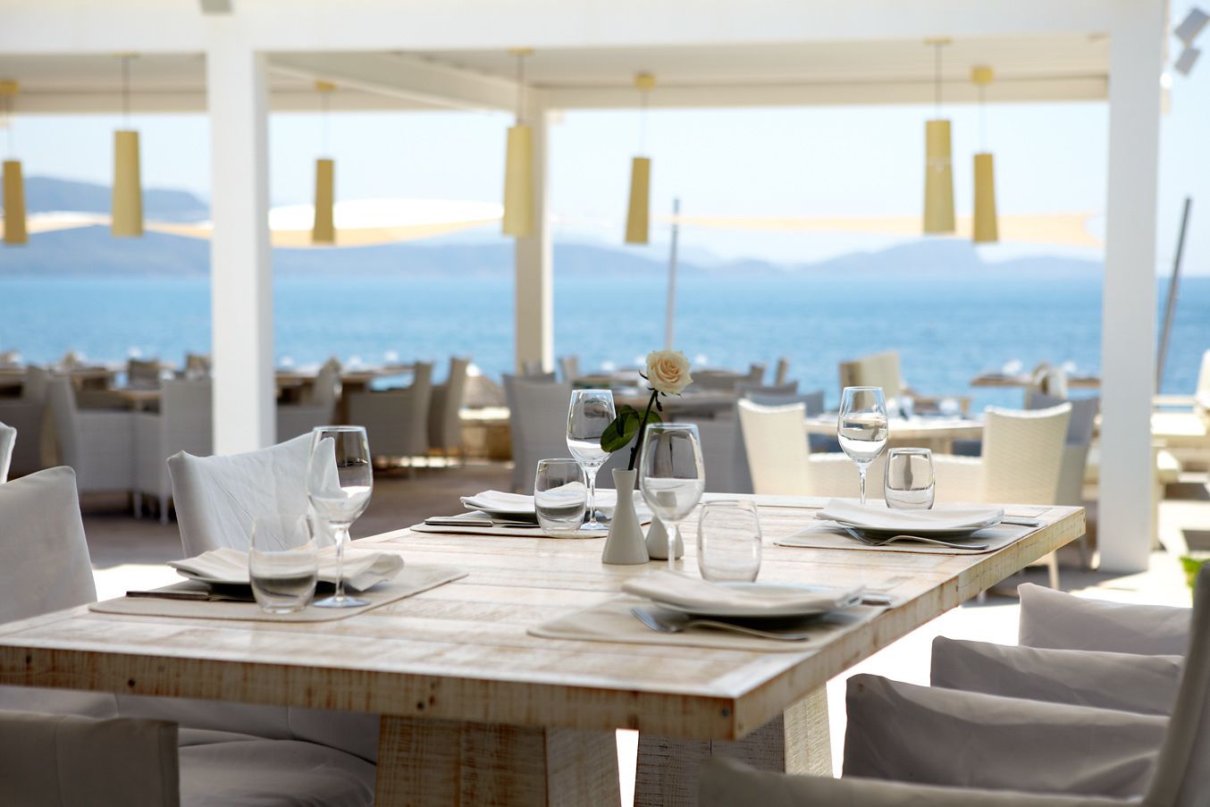 A restaurant with a wooden table set for dining, white chairs, and a view of the sea.