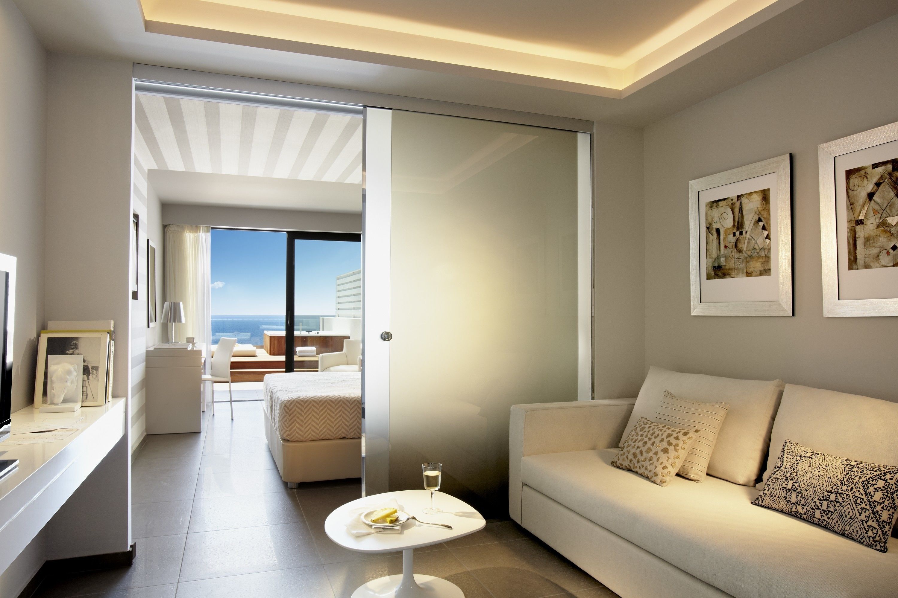 A modern hotel room with a sofa, a small table with a glass of water, and a view of the sea through a sliding door.