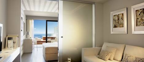 A modern hotel room with a sofa, a small table with a glass of water, and a view of the sea through a sliding door.