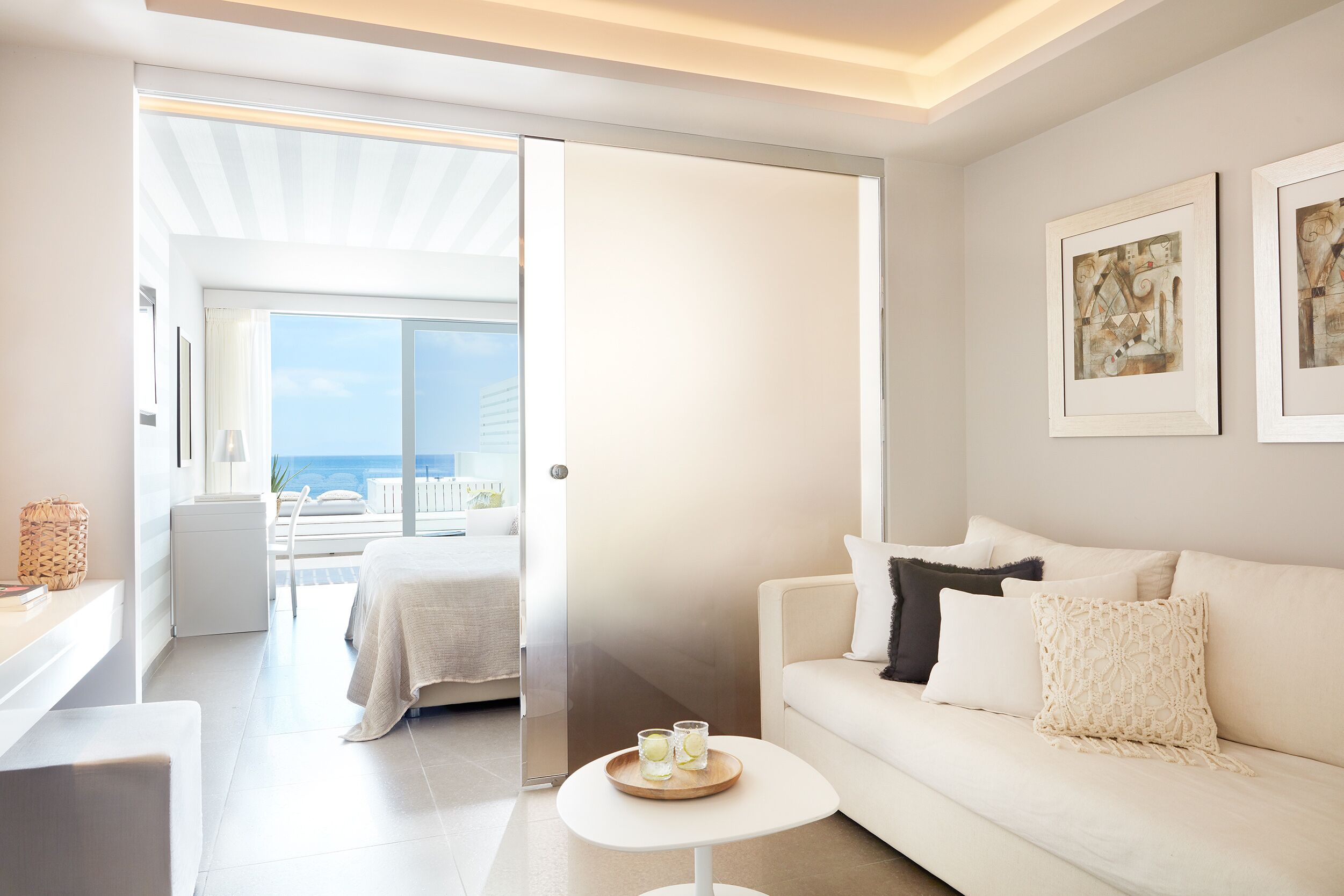 Habitación de hotel moderna con un sofá blanco, una mesita redonda con vasos y un balcón con vista al mar.