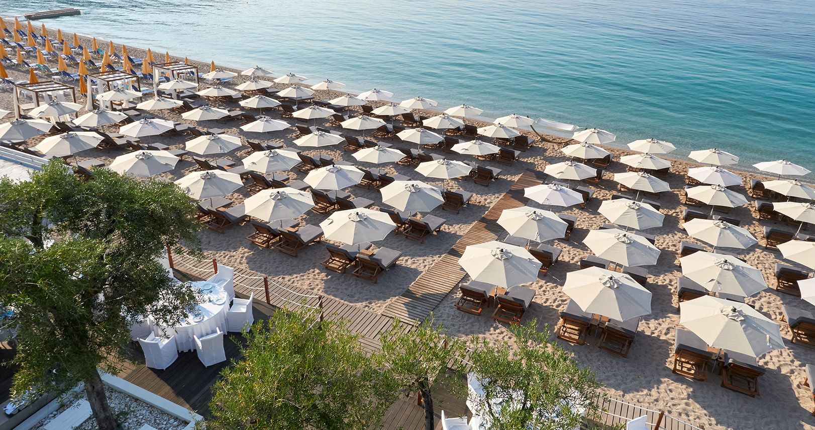 An aerial view of a beachside resort with rows of sun umbrellas and lounge chairs, a wooden deck area, and a pool.
