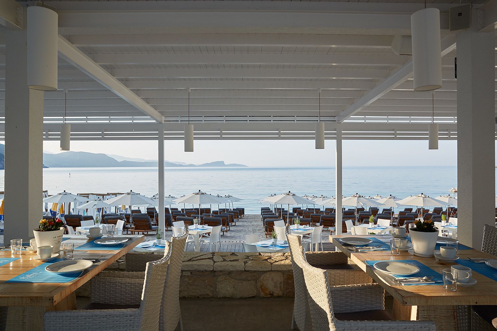 Ein Restaurant am Meer mit Außenbereich und Meerblick, mit Tischen unter Sonnenschirmen und Korbstühlen.