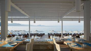 Ein Restaurant am Meer mit Außenbereich und Meerblick, mit Tischen unter Sonnenschirmen und Korbstühlen.