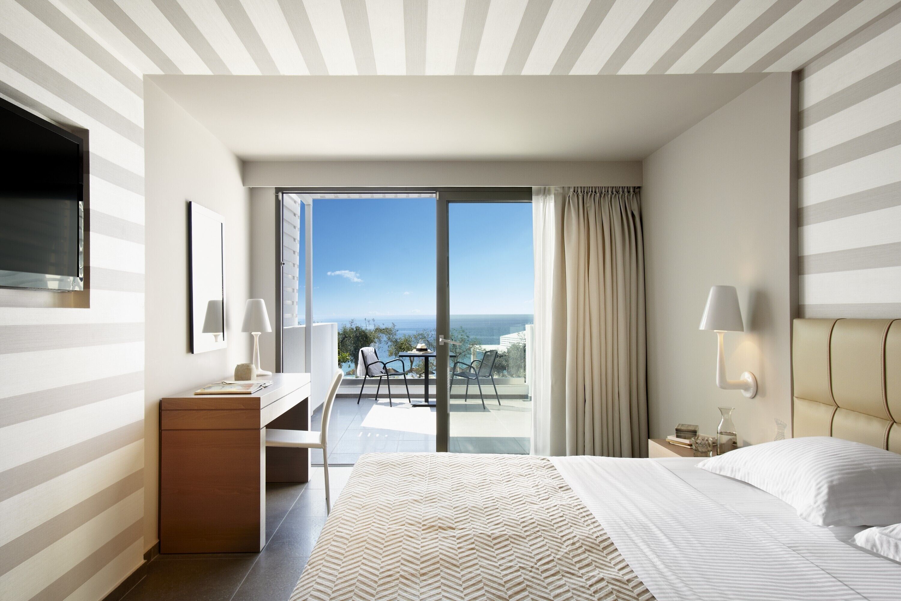 A hotel room with a bed, a desk, a chair, a TV, a balcony with a view of the sea, and a window with curtains.