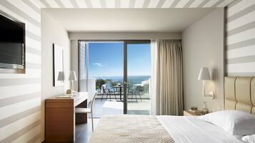 A hotel room with a bed, a desk, a chair, a TV, a balcony with a view of the sea, and a window with curtains.