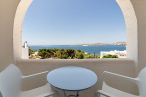 A balcony with a table and chairs, overlooking a coastal view with buildings and trees. - Stratos Studios & Apartments (Paros)